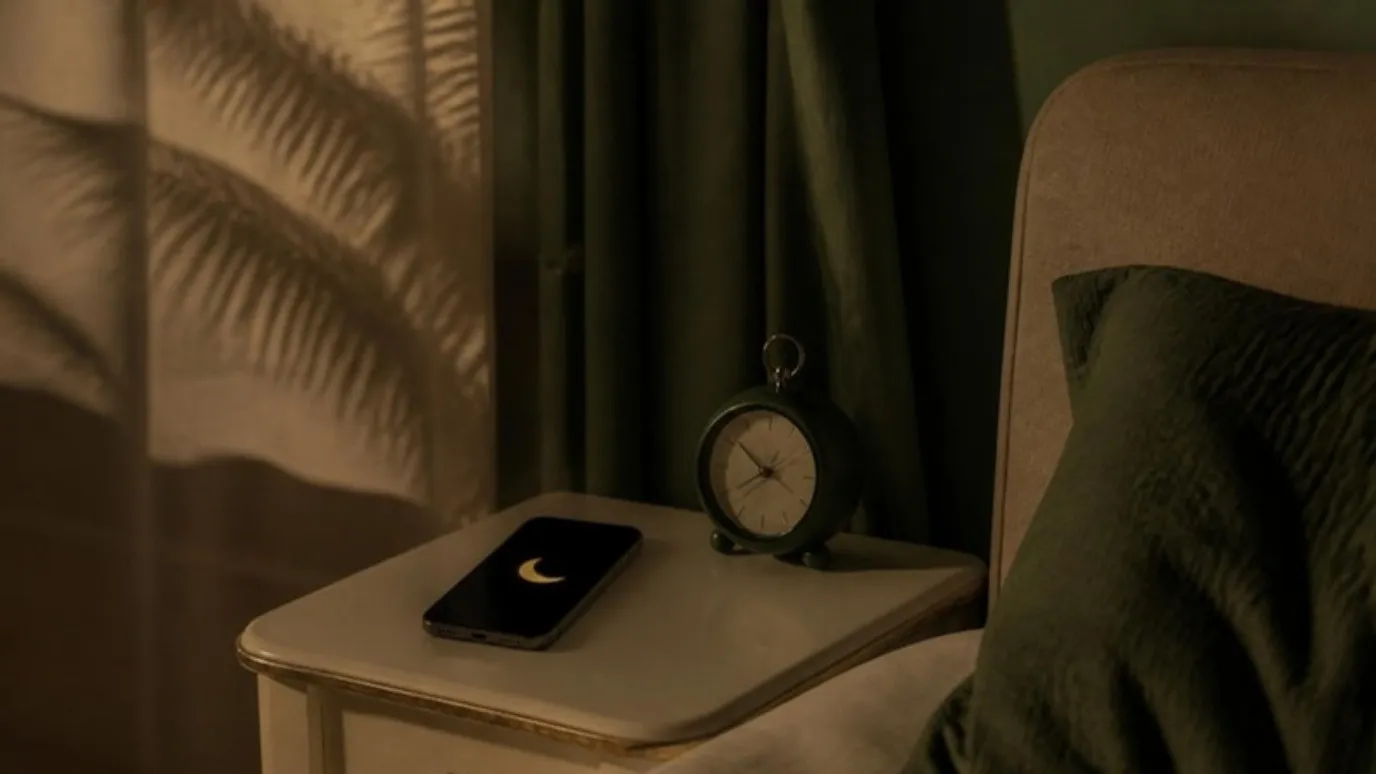 Un teléfono con el ícono de una luna en la mesa de noche, junto a un reloj pequeño, en un cuarto tranquilo de noche.