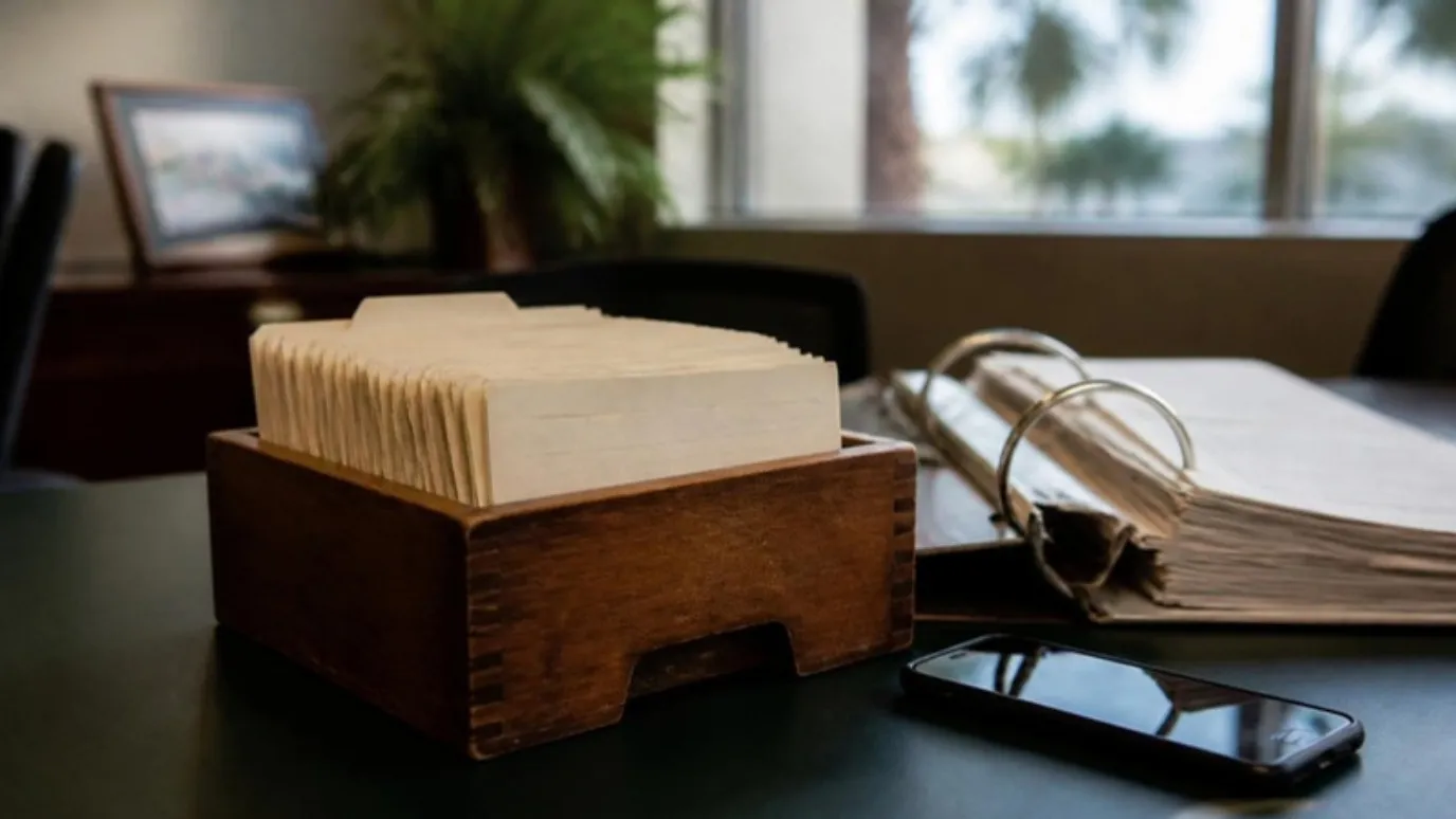 Una caja de madera con fichas de clientes en un escritorio, junto a una carpeta de trabajos viejos y un teléfono.