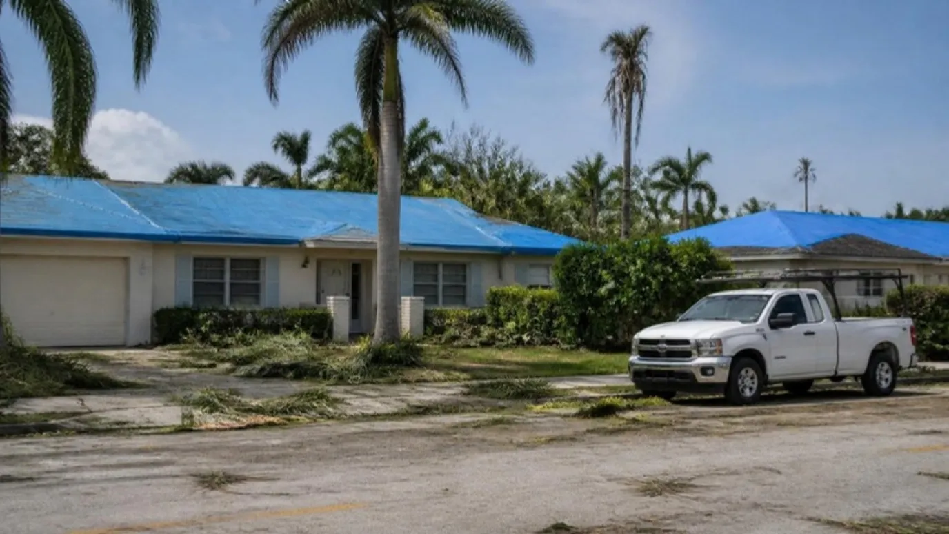 Una calle después de una tormenta con techos cubiertos de azul, pencas de palma en el suelo y una camioneta blanca en la orilla.